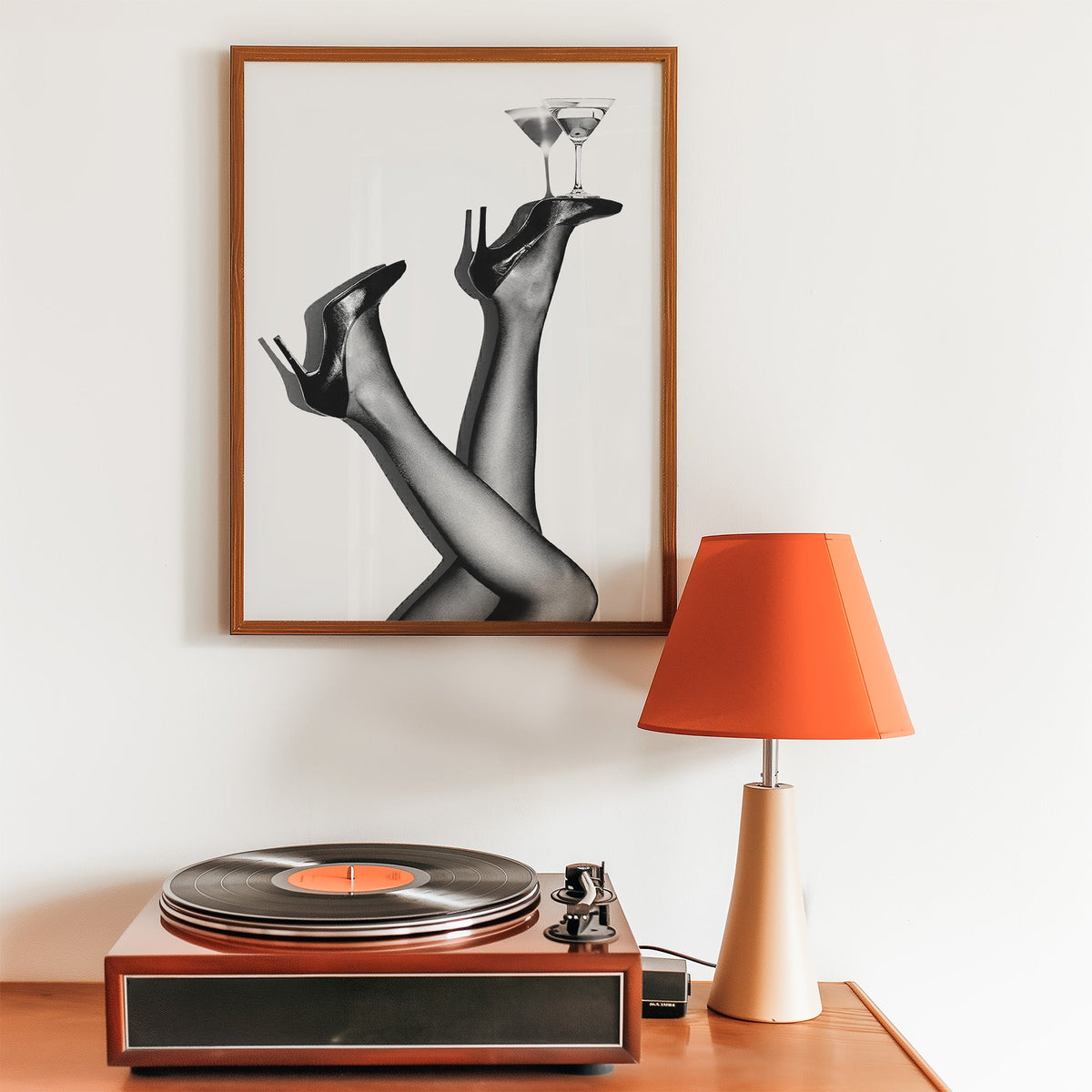 Legs in heels balancing two martini glasses print in a walnut frame above a record player and orange lamp