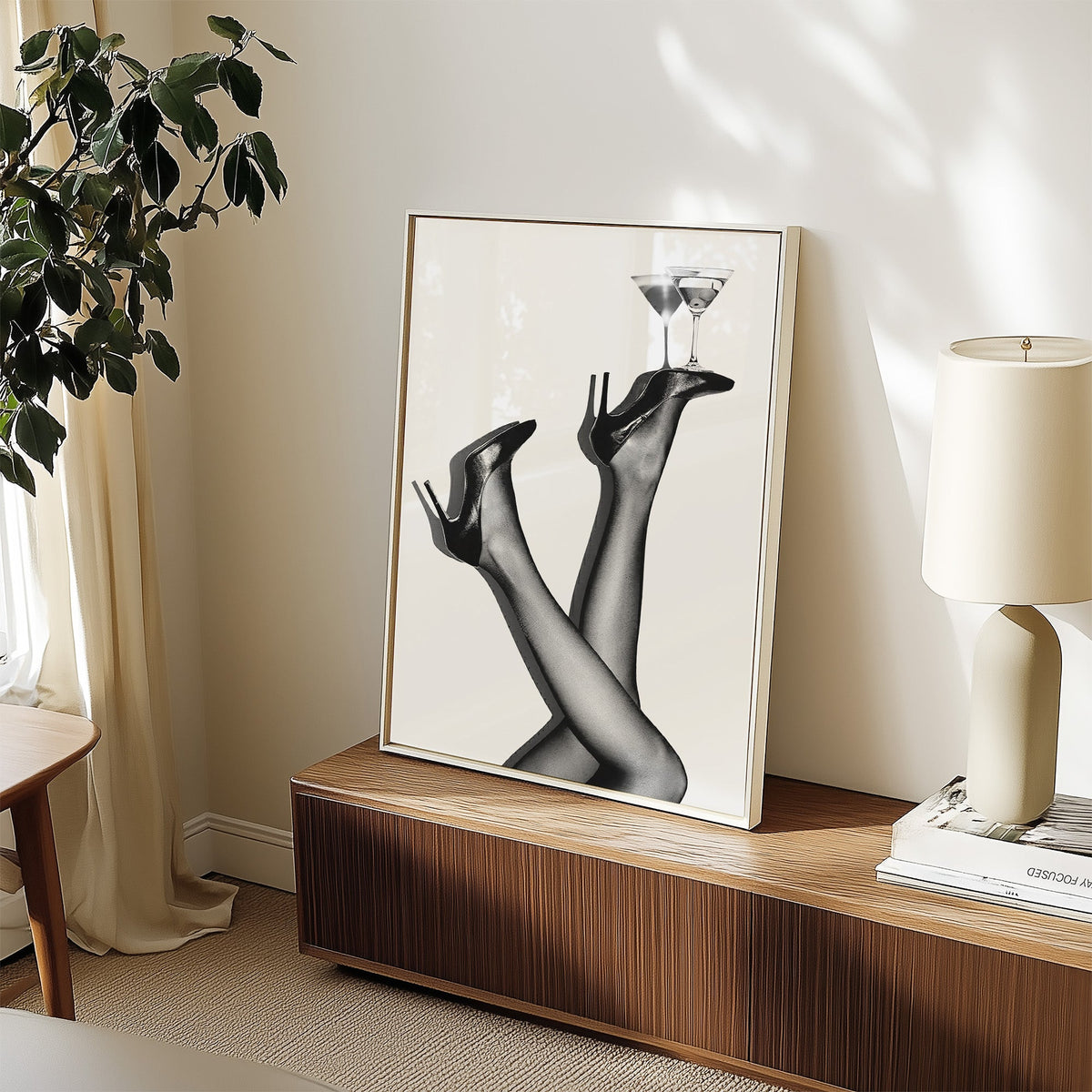 Legs in heels balancing two martini glasses print in a white frame on a walnut sideboard beside a ceramic lamp