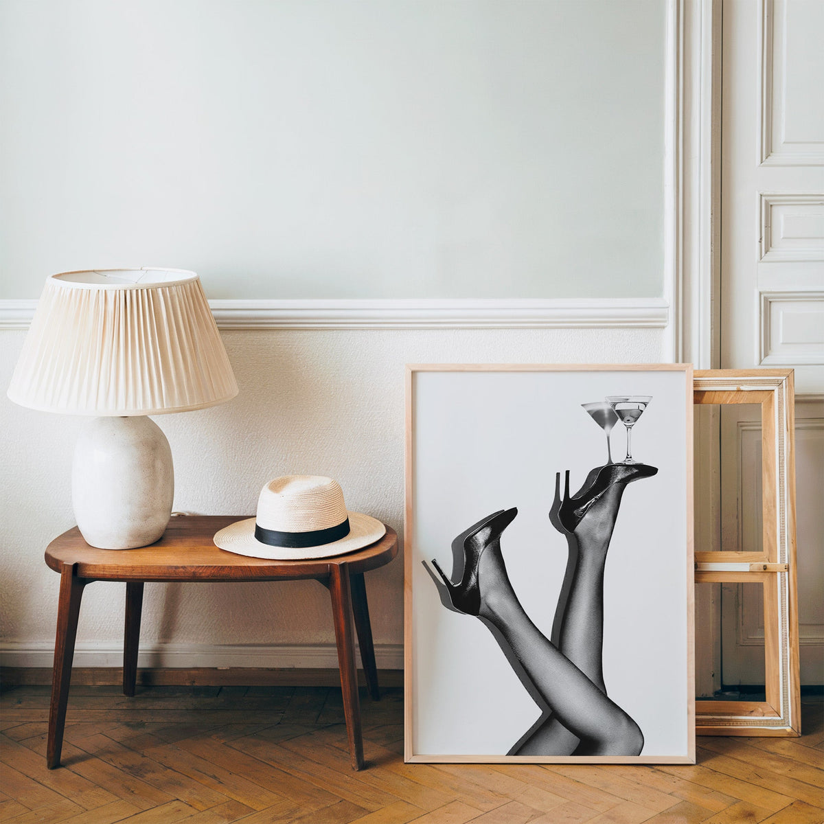 Legs in heels balancing two martini glasses print in a pine frame on a wood floor beside a lamp and straw hat