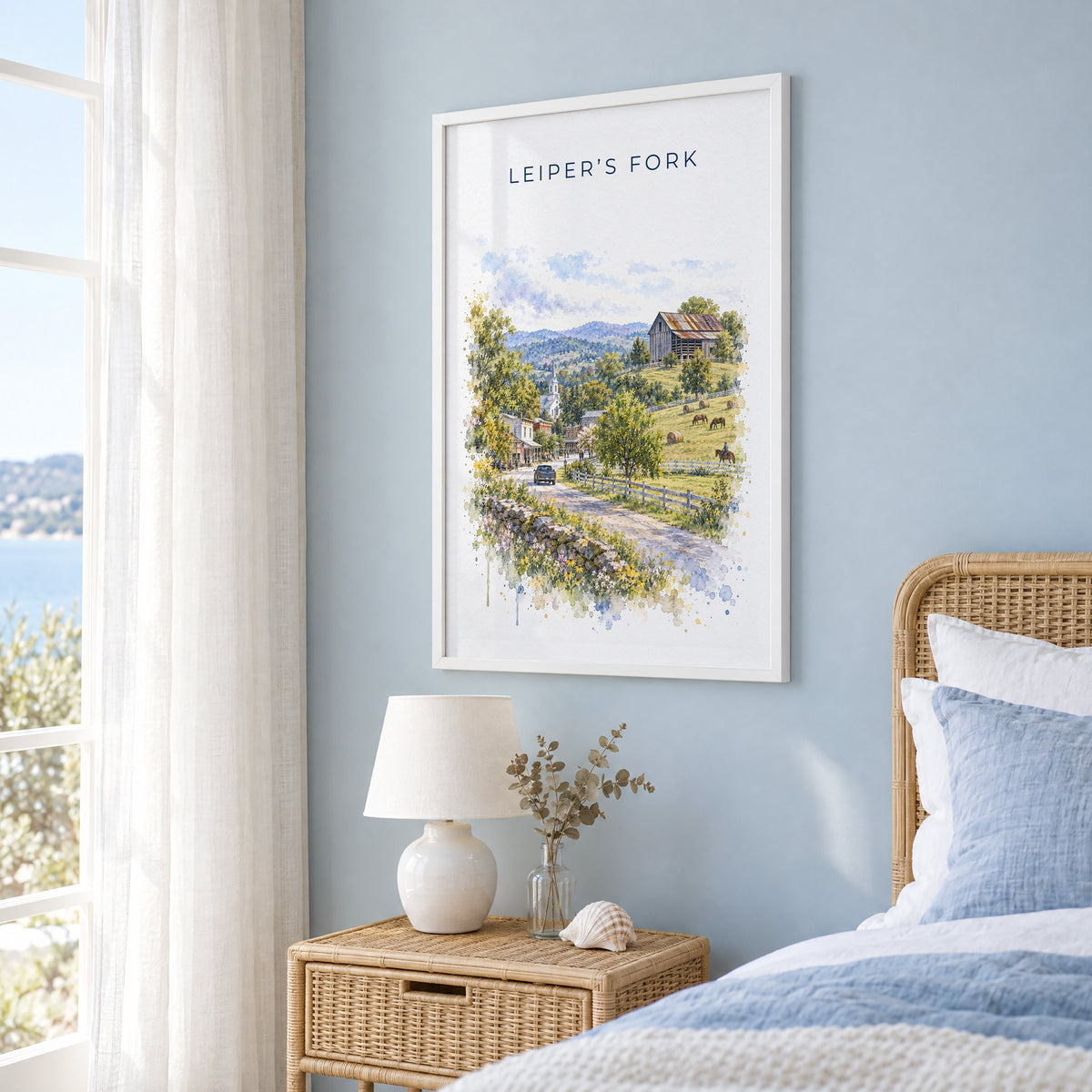 Leiper's Fork, Tennessee watercolor print in a white frame on a pale blue wall beside a rattan bed in a coastal bedroom