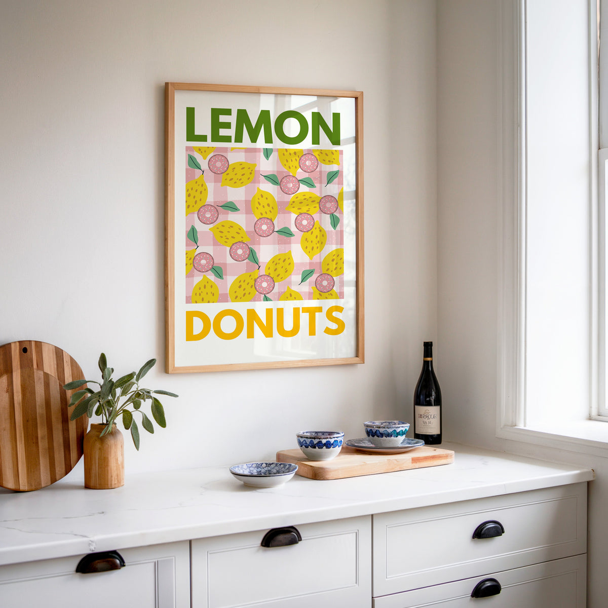Lemon and donuts print in an oak frame above a white sideboard with a bottle of wine