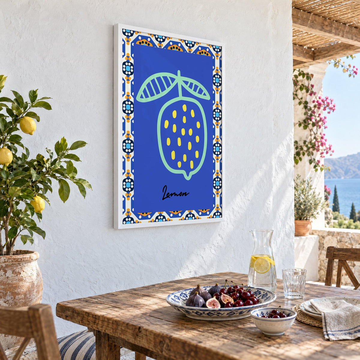 Lemon mint outline cobalt tile print in a white frame on a whitewashed terrace wall above a lunch table with figs and cherries, seen at an angle