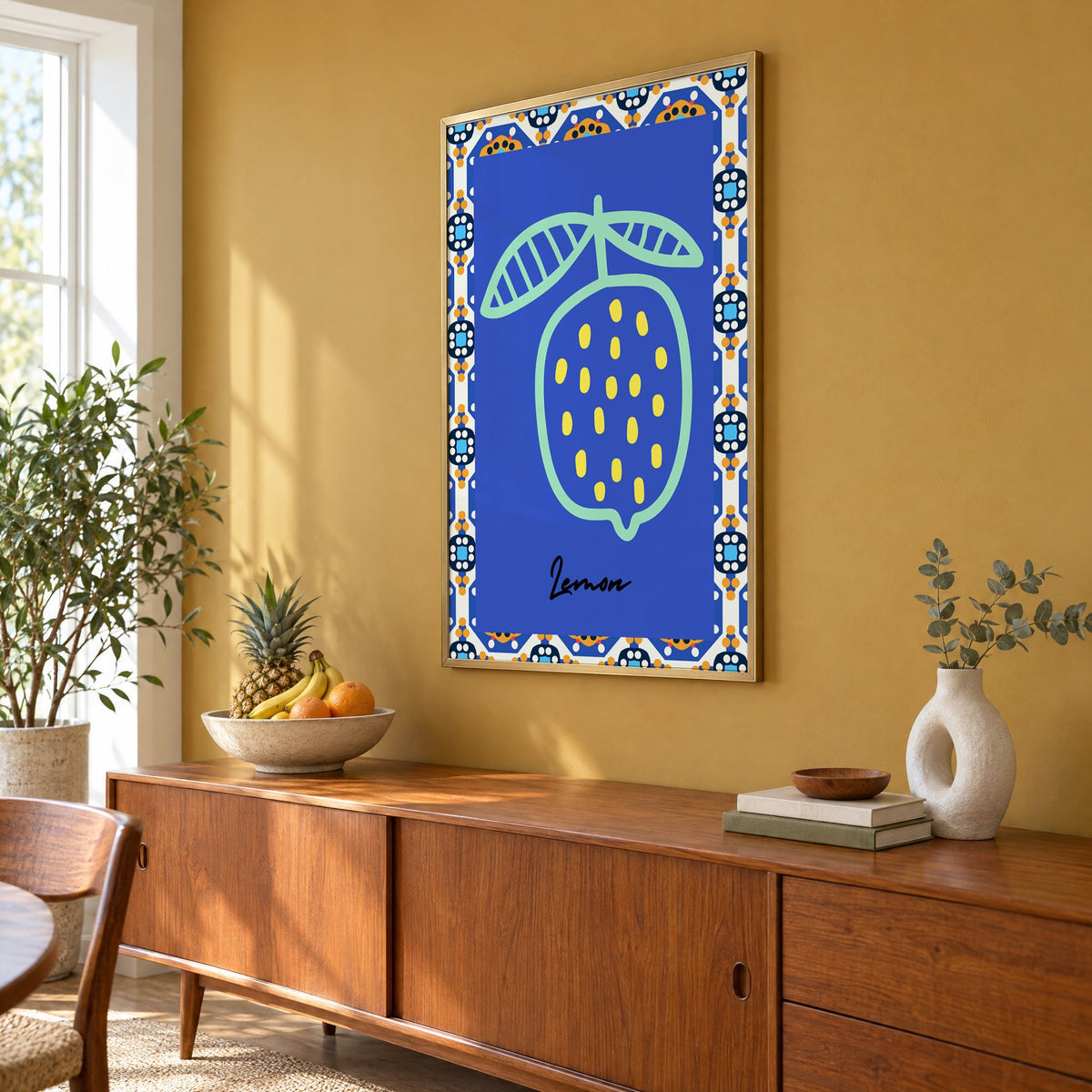 Lemon mint outline cobalt tile print in a brass frame on a mustard wall above a teak sideboard with a fruit bowl, seen at an angle