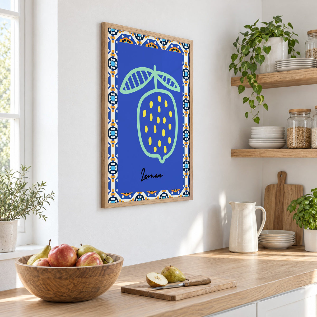 Lemon mint outline cobalt tile print in an oak frame on a white kitchen wall above a counter with apples and pears, seen at an angle