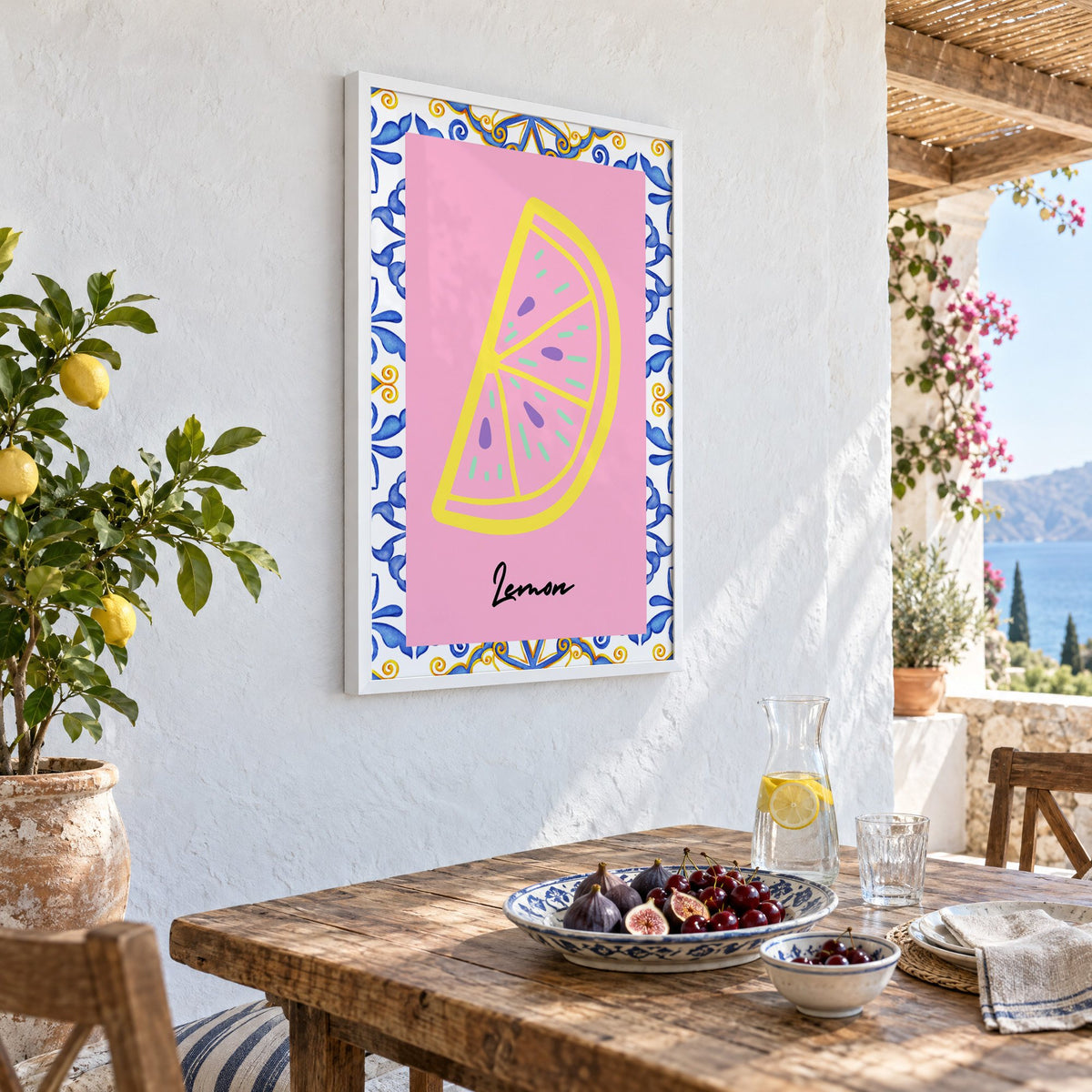 Lemon slice pink majolica tile print in a white frame on a whitewashed terrace wall above a lunch table with figs and cherries, seen at an angle