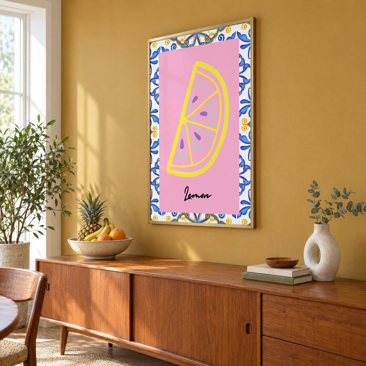 Lemon slice pink majolica tile print in a brass frame on a mustard wall above a teak sideboard with a fruit bowl, seen at an angle