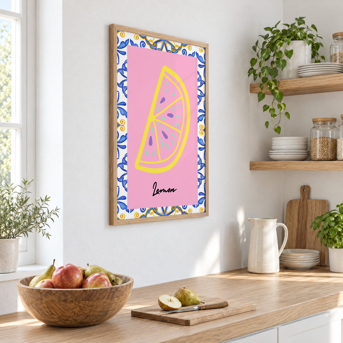 Lemon slice pink majolica tile print in an oak frame on a white kitchen wall above a counter with apples and pears, seen at an angle