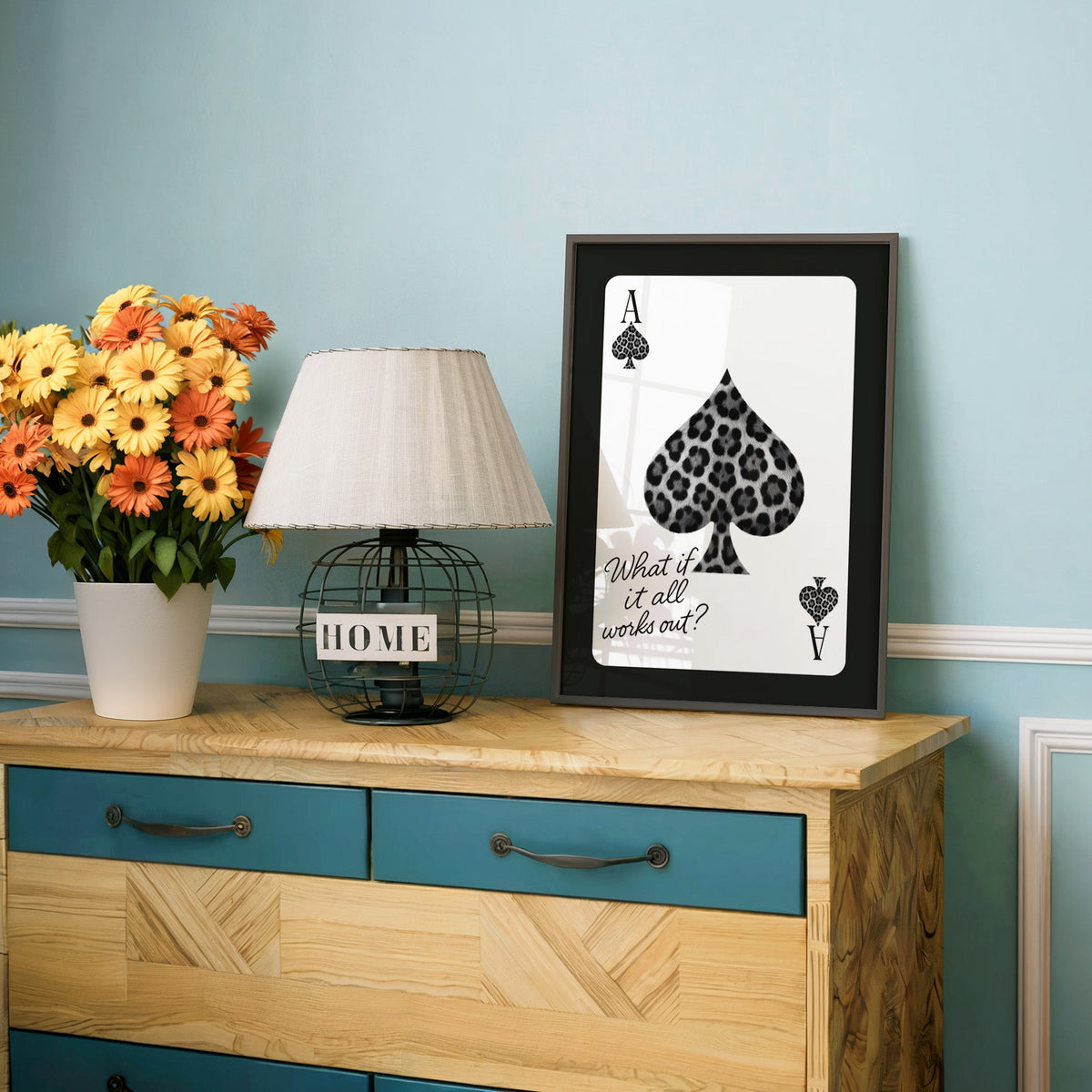 Leopard fur ace of spades card print in a charcoal frame on a teal dresser beside orange daisies