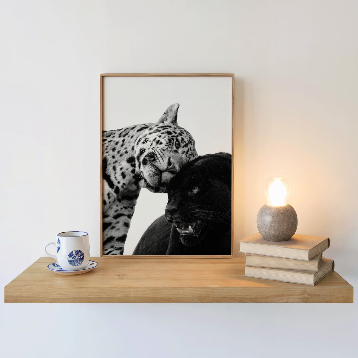 Leopard nuzzling black panther close-up print in an oak frame on a floating shelf beside a glowing lamp