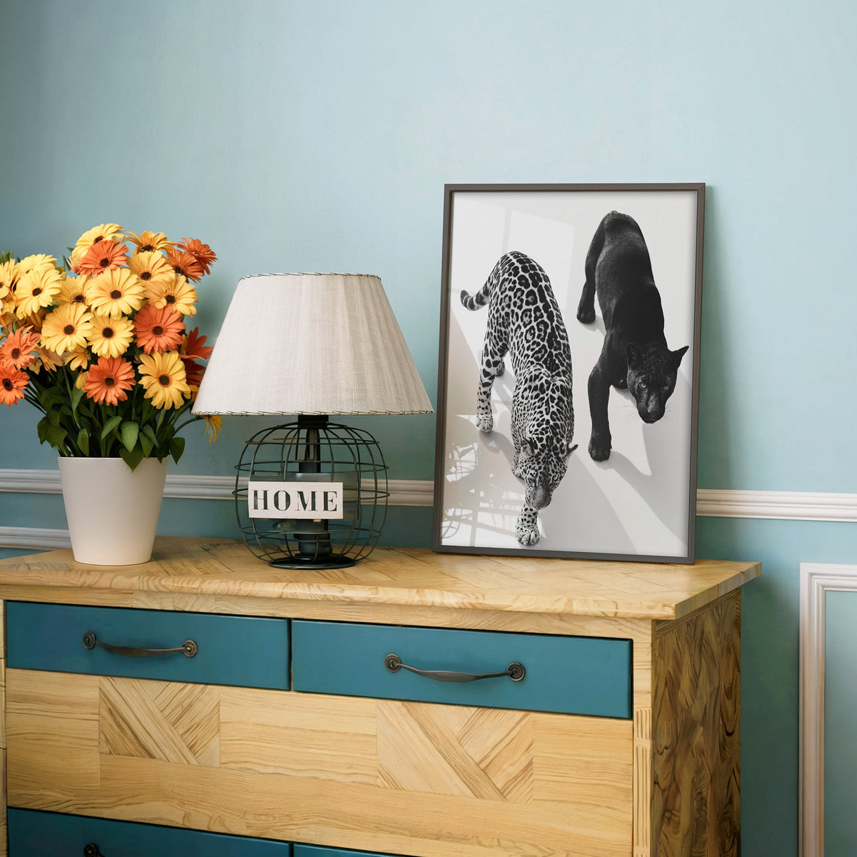 Overhead view of leopard and black panther print in a charcoal frame on a teal dresser beside orange daisies