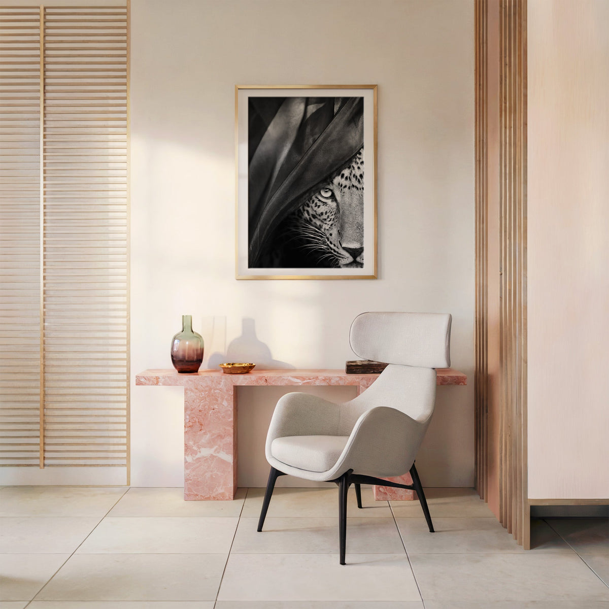 Leopard eye peering through black veil print shown in a gold frame above a pink marble console and armchair
