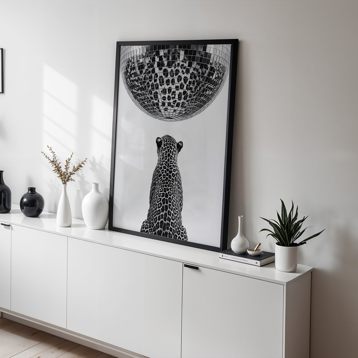 Leopard looking up at giant disco ball print in a black frame on a white sideboard with black and white vases