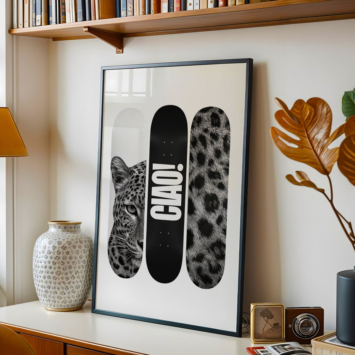 Three skateboard decks with leopard face and CIAO print in a black frame on a console under a bookshelf
