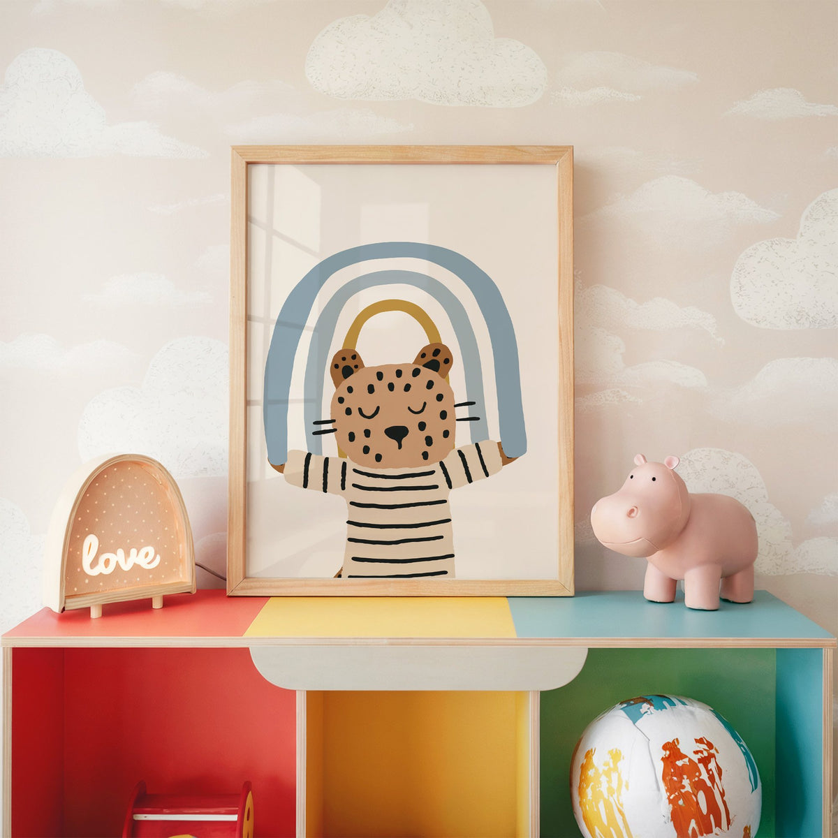 Leopard holding blue rainbow nursery print in an oak frame on a rainbow toy shelf against cloud wallpaper