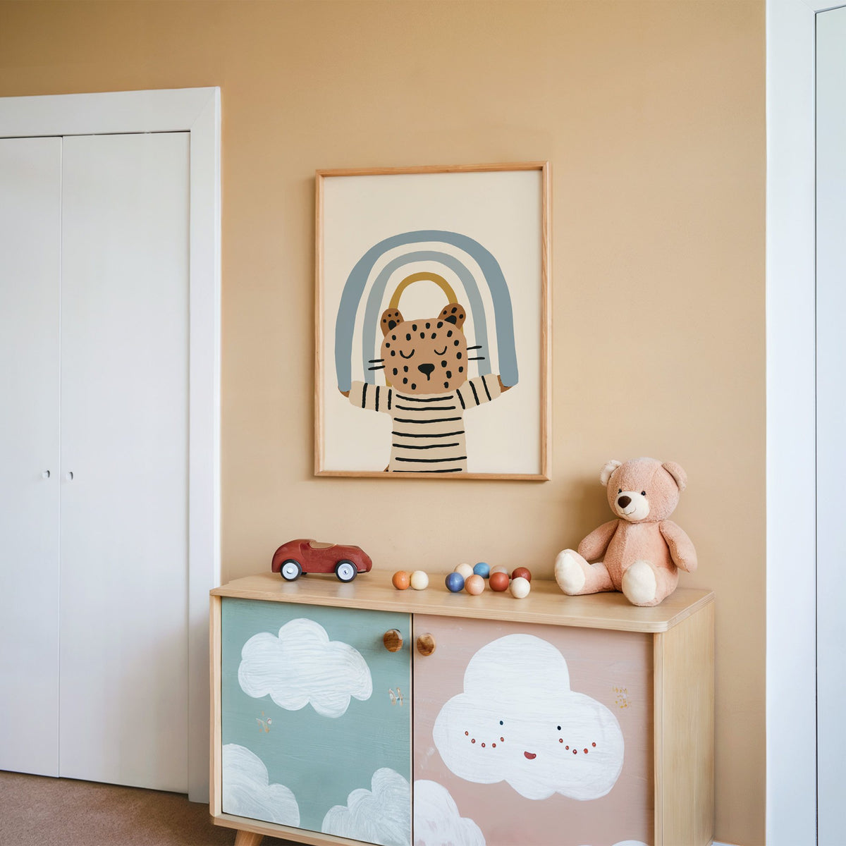 Leopard holding blue rainbow nursery print in an oak frame on a peach wall above a cloud-painted cabinet
