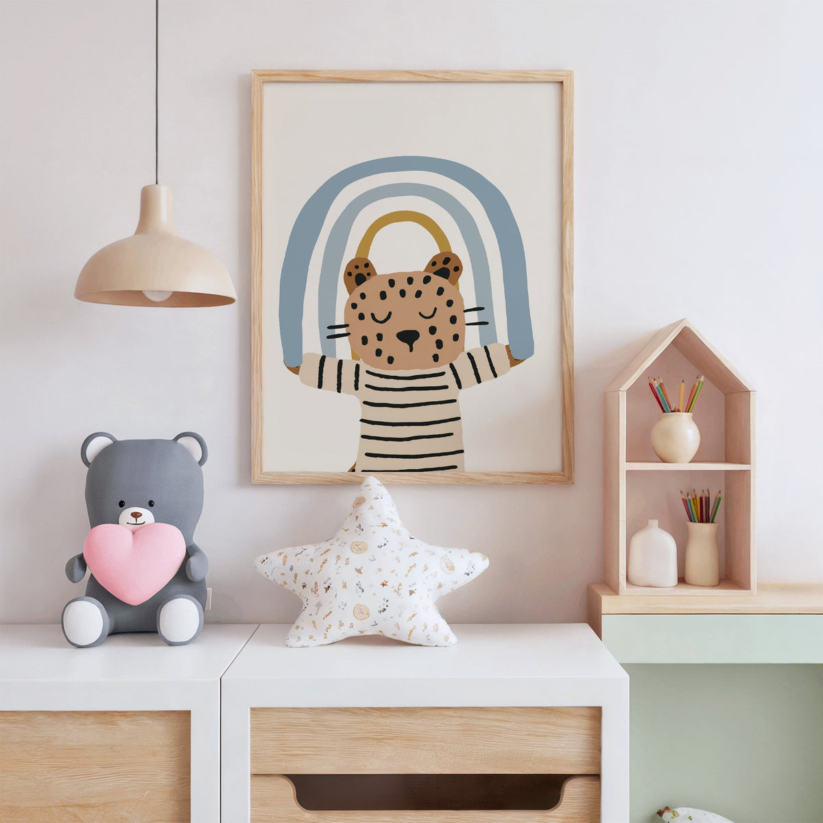 Leopard holding blue rainbow nursery print in an oak frame above a white dresser with a star pillow and house shelf