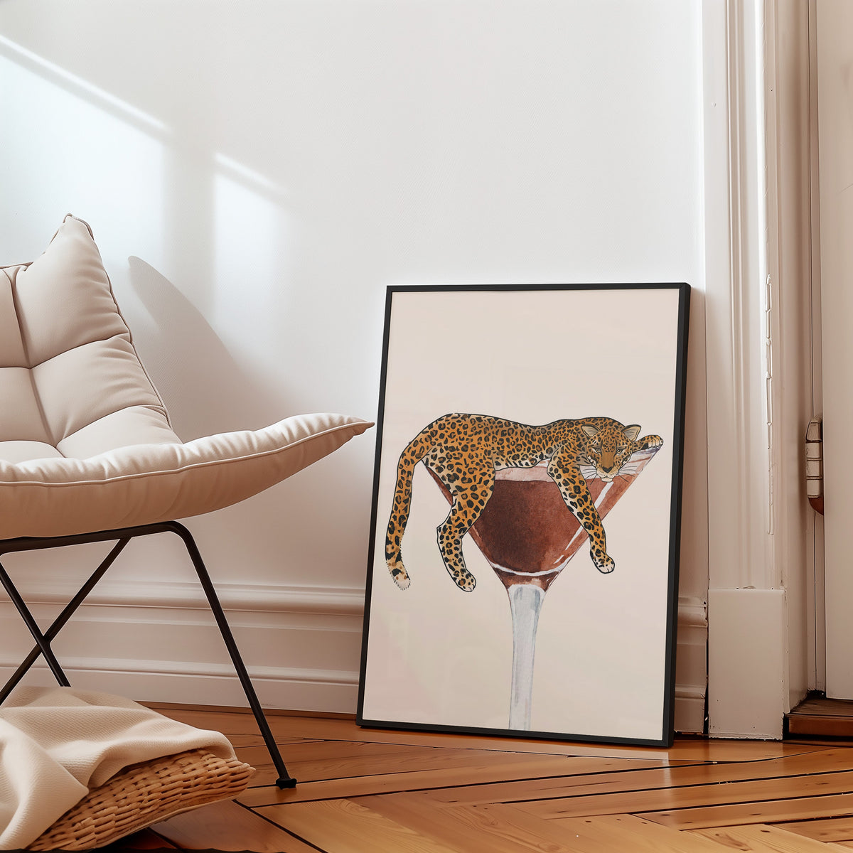 Leopard draped on a martini glass print in a slim black frame leaning on a herringbone floor beside a cream armchair