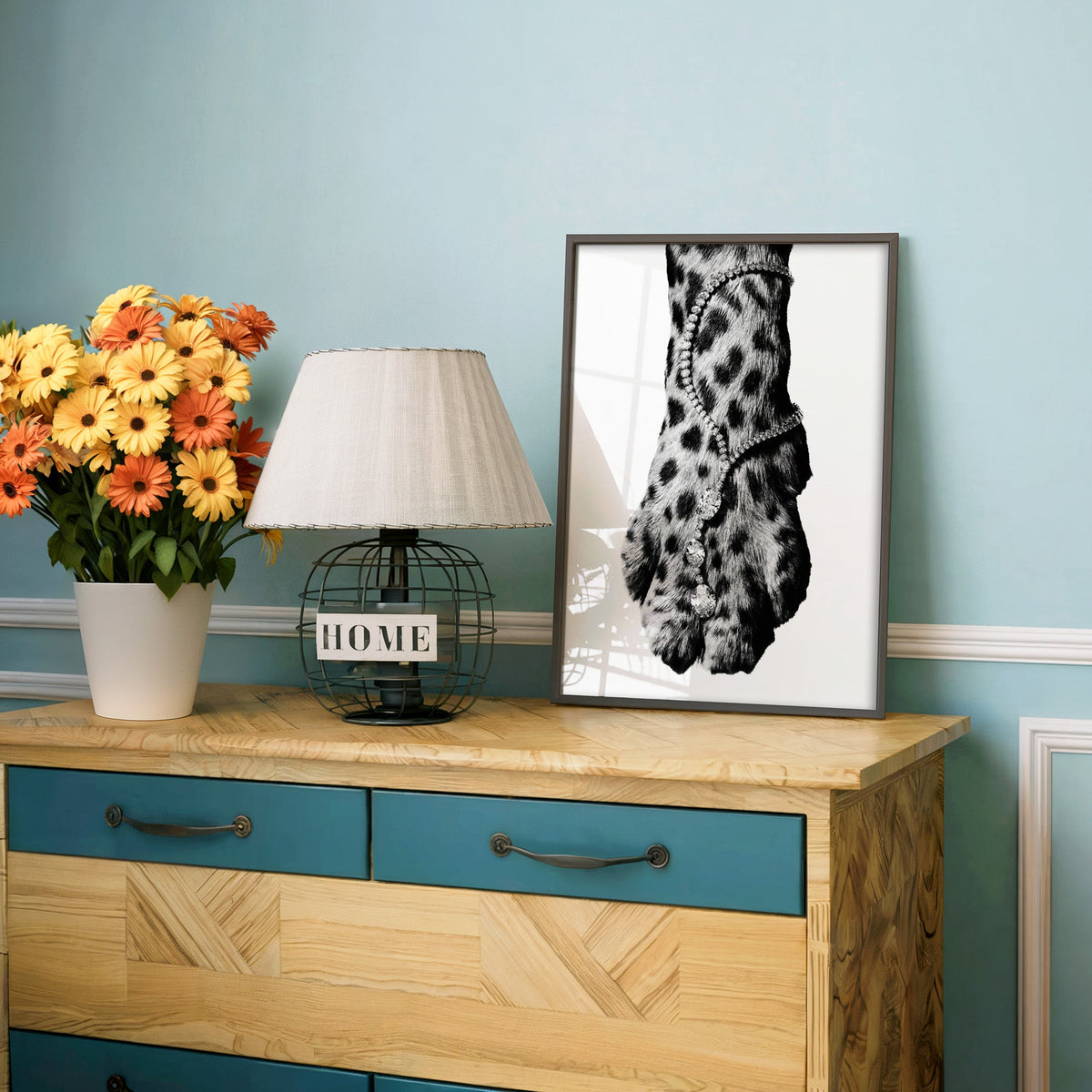 Leopard paw with sparkling stone anklet close-up print in a charcoal frame on a teal dresser beside orange daisies