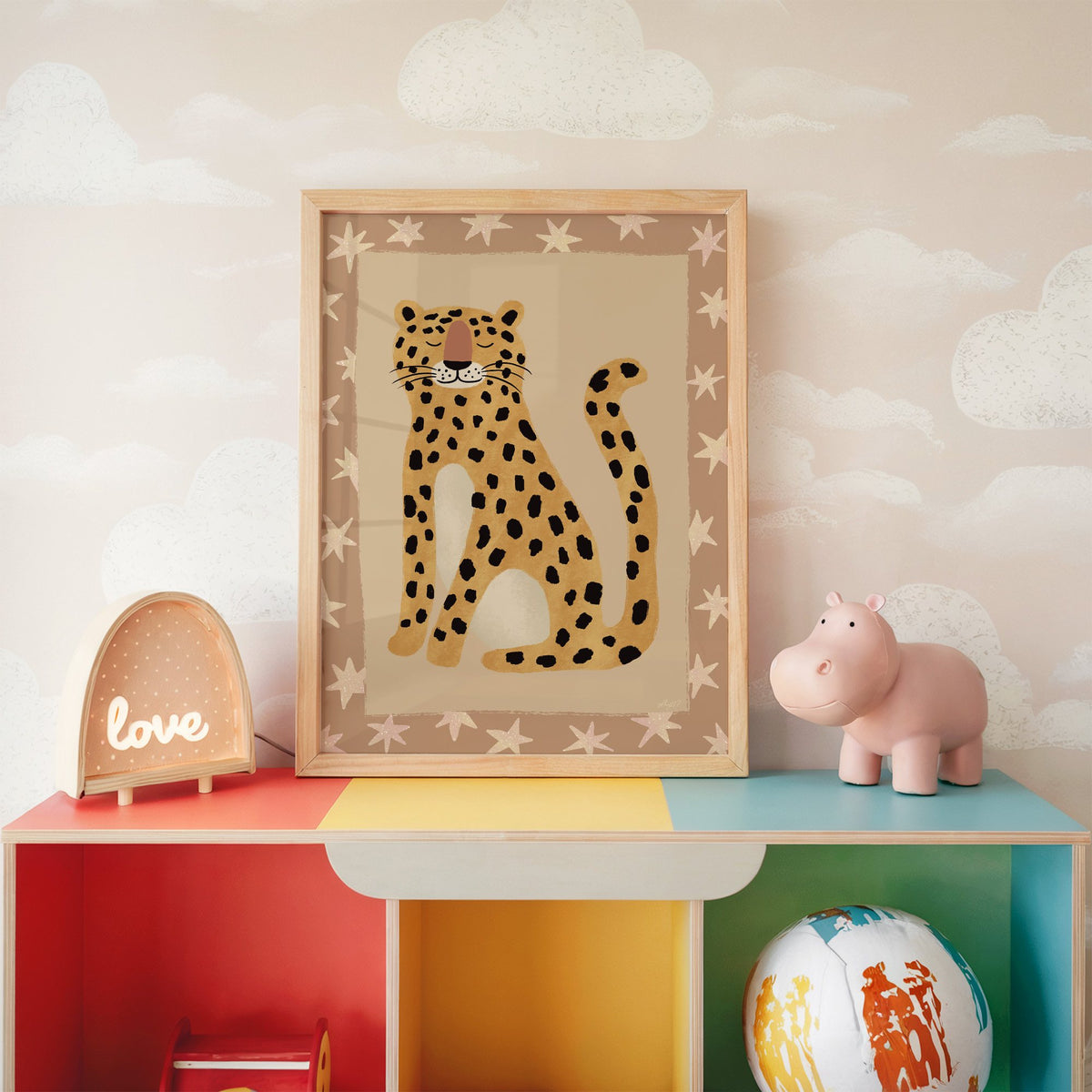 Smiling leopard with star border nursery print in an oak frame on a rainbow toy shelf against cloud wallpaper