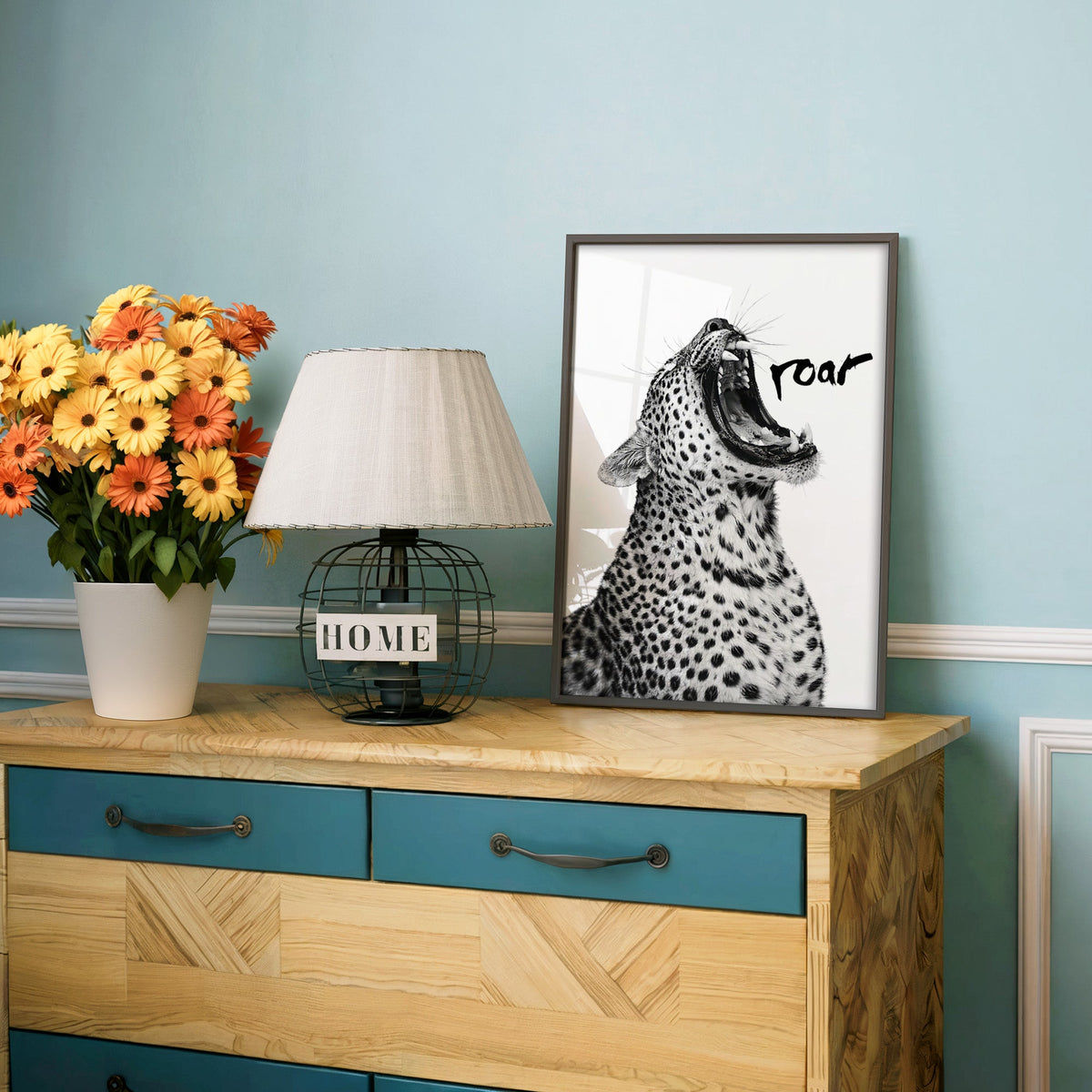Roaring leopard with black brush lettering print in a charcoal frame on a teal dresser beside orange daisies