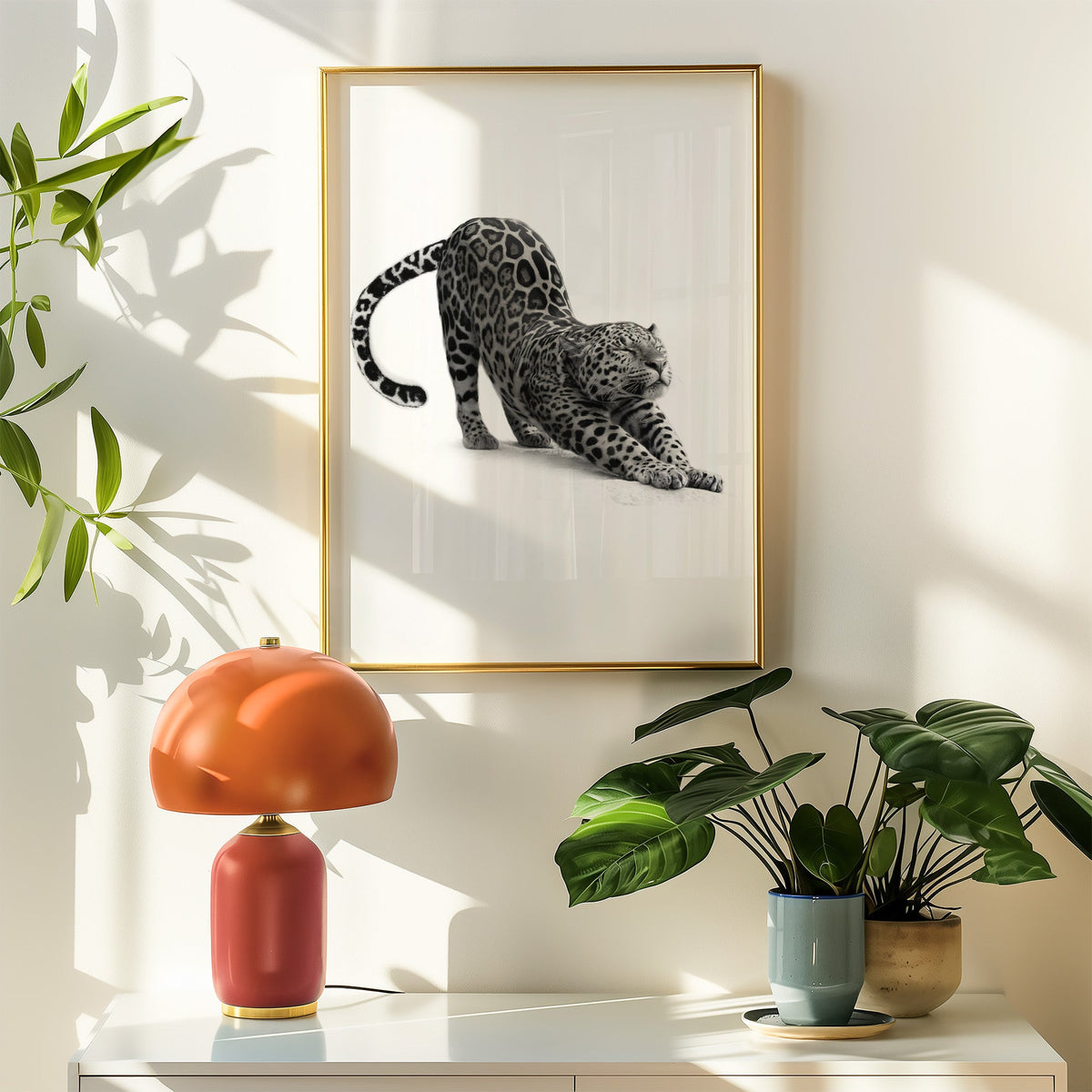 Stretching leopard with curled tail print shown in a gold frame above a white sideboard with an orange lamp