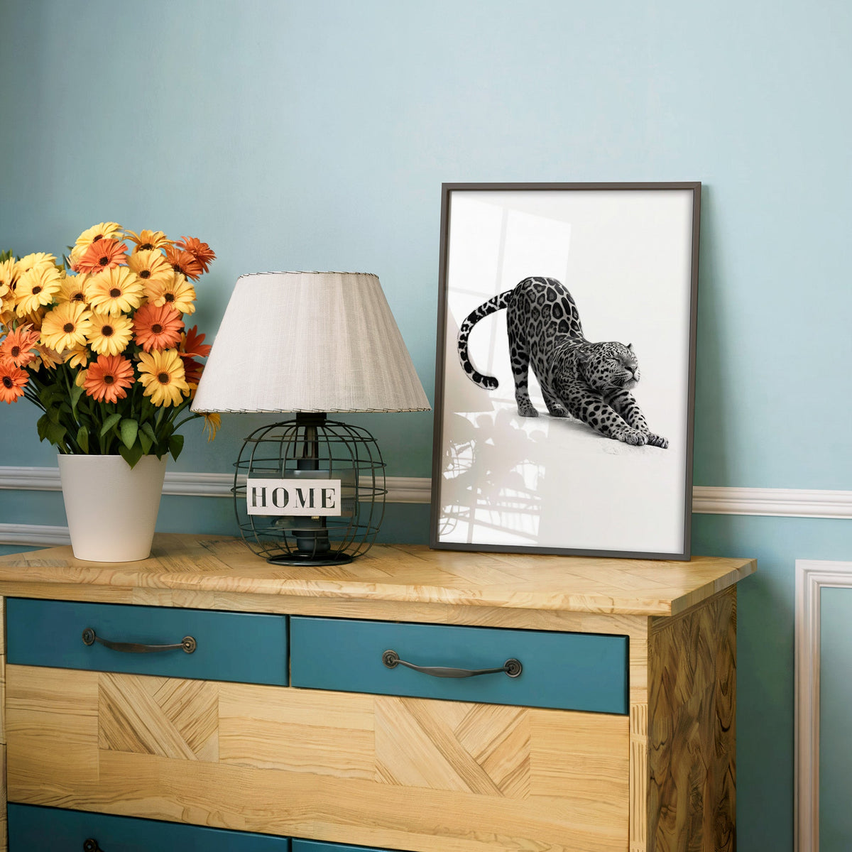 Stretching leopard with curled tail print in a charcoal frame on a teal dresser beside orange daisies