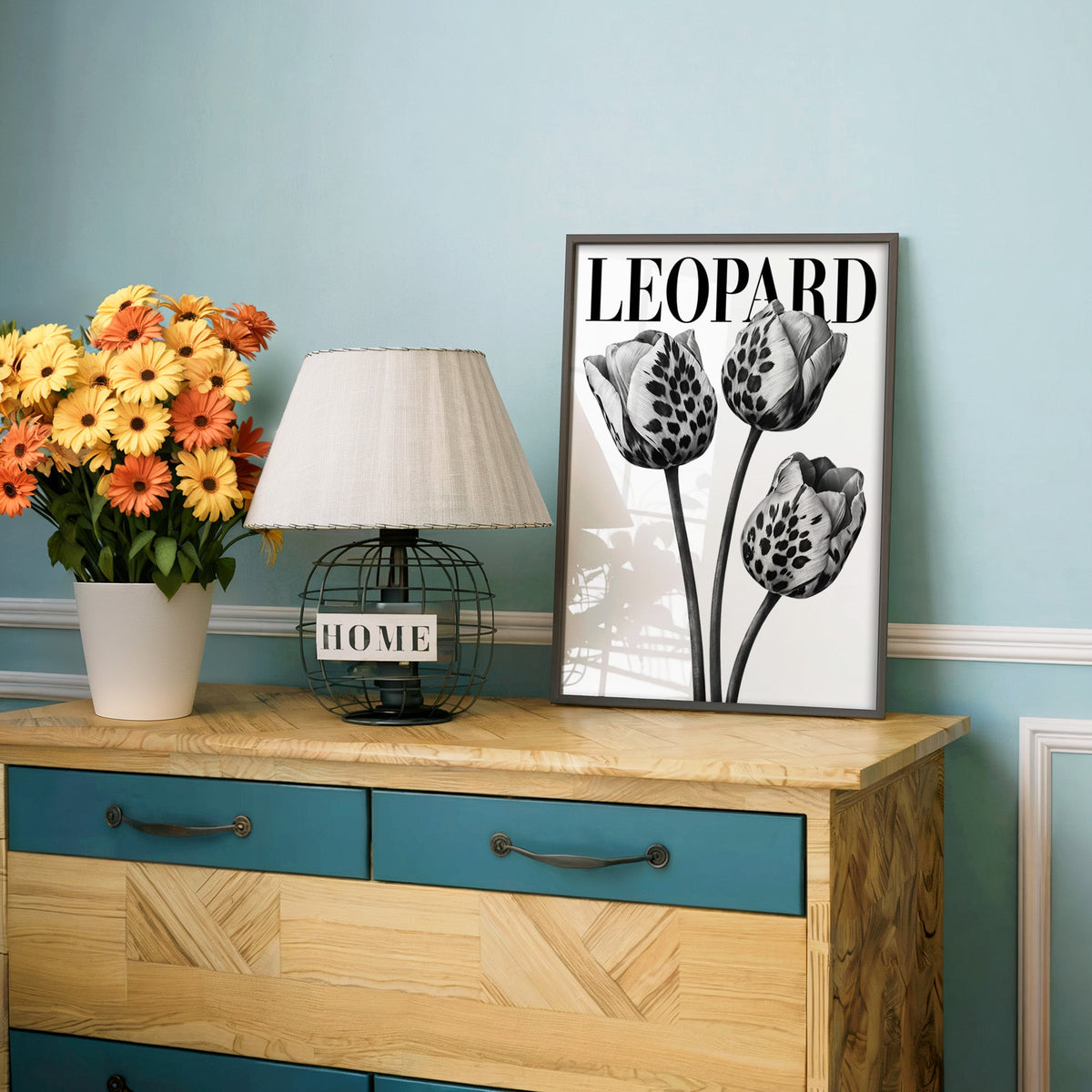 Three leopard-spotted tulips with LEOPARD title print in a charcoal frame on a teal dresser beside orange daisies