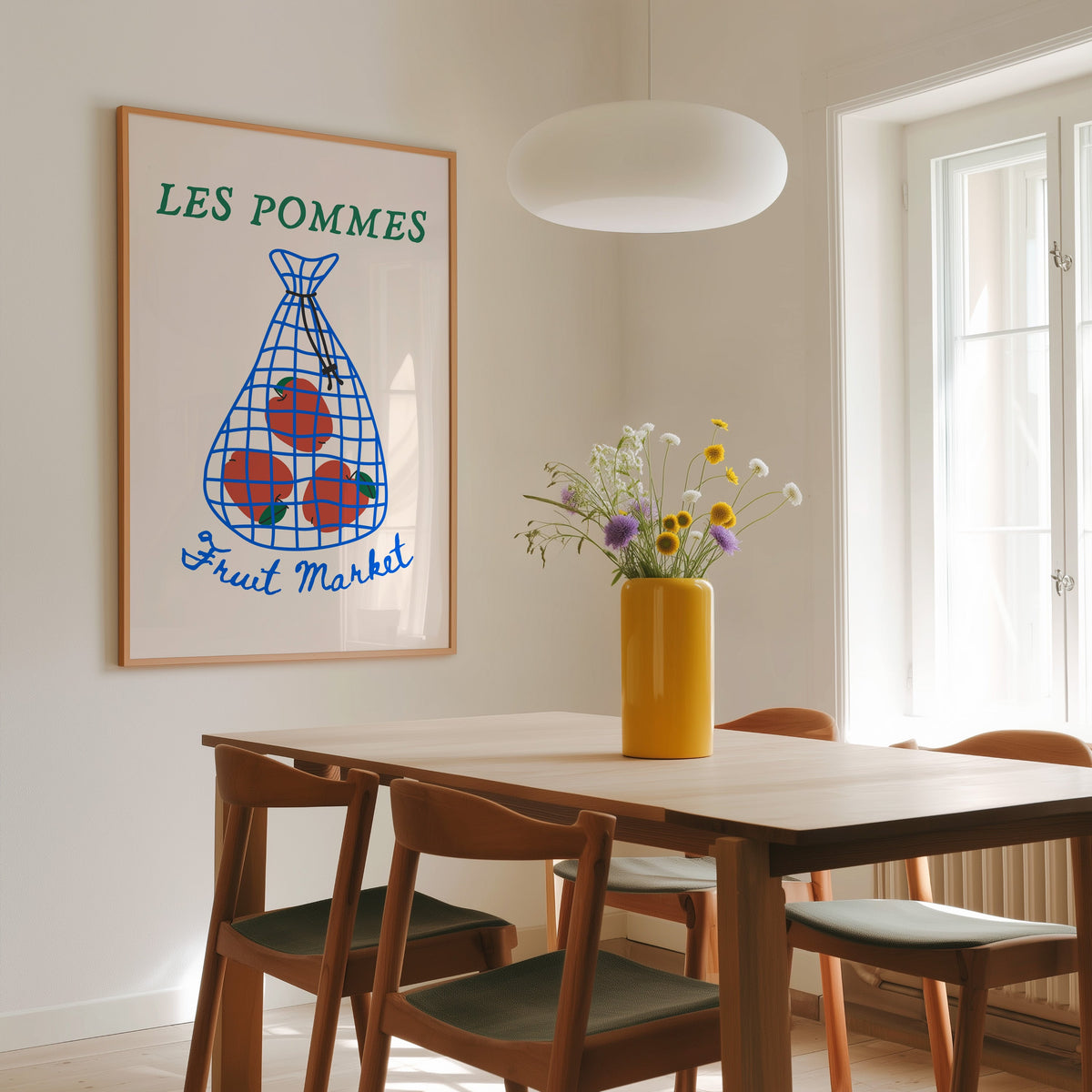 Les Pommes apples in a net bag print in a slim oak frame above a wooden dining table under a round pendant light