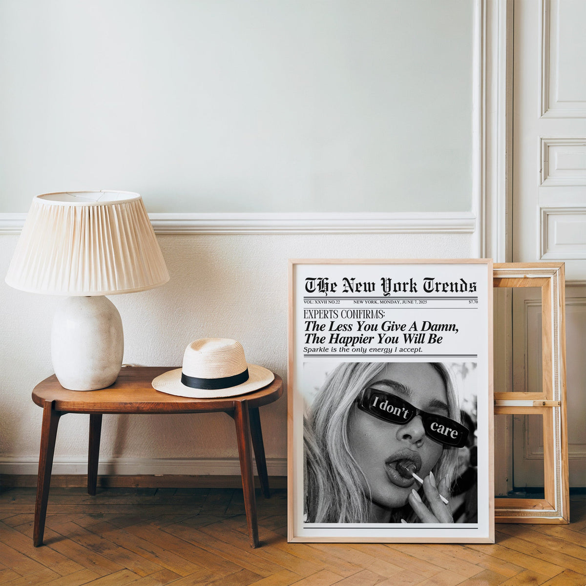 I don't care glasses and lollipop mock newspaper print in a pine frame on a wood floor beside a lamp and straw hat