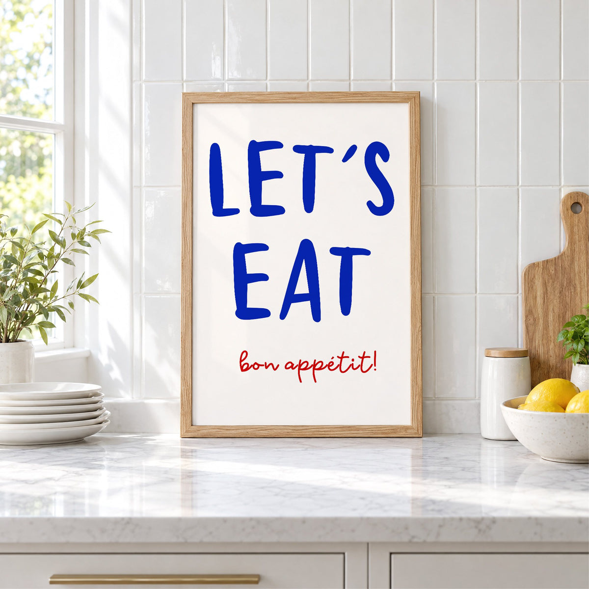 Let's Eat cobalt blue type print in an oak frame leaning on a marble counter by white tiles and lemons