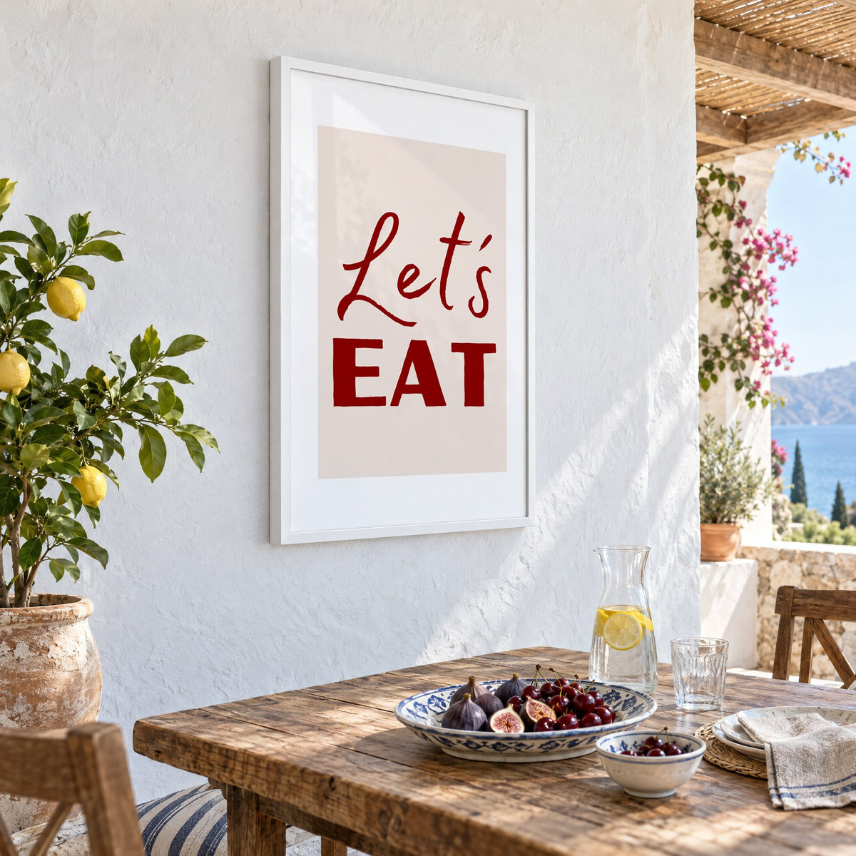 Let's EAT burgundy script print in a white frame on a whitewashed terrace wall above a lunch table with figs and cherries, seen at an angle