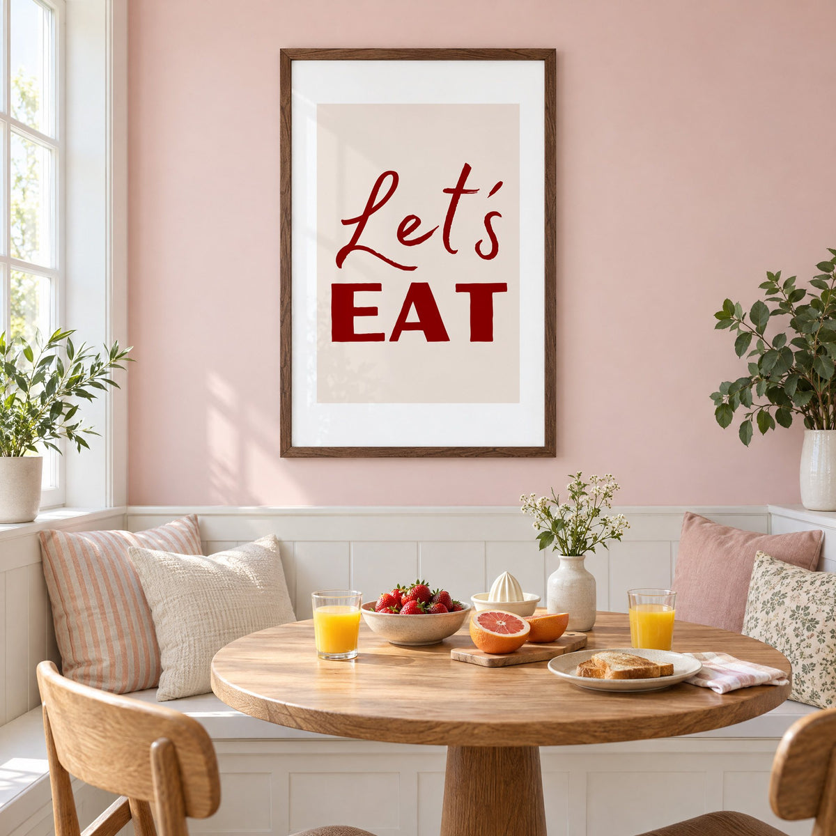 Let's EAT burgundy script print in a walnut frame on a pastel pink wall above a breakfast nook with strawberries and orange juice