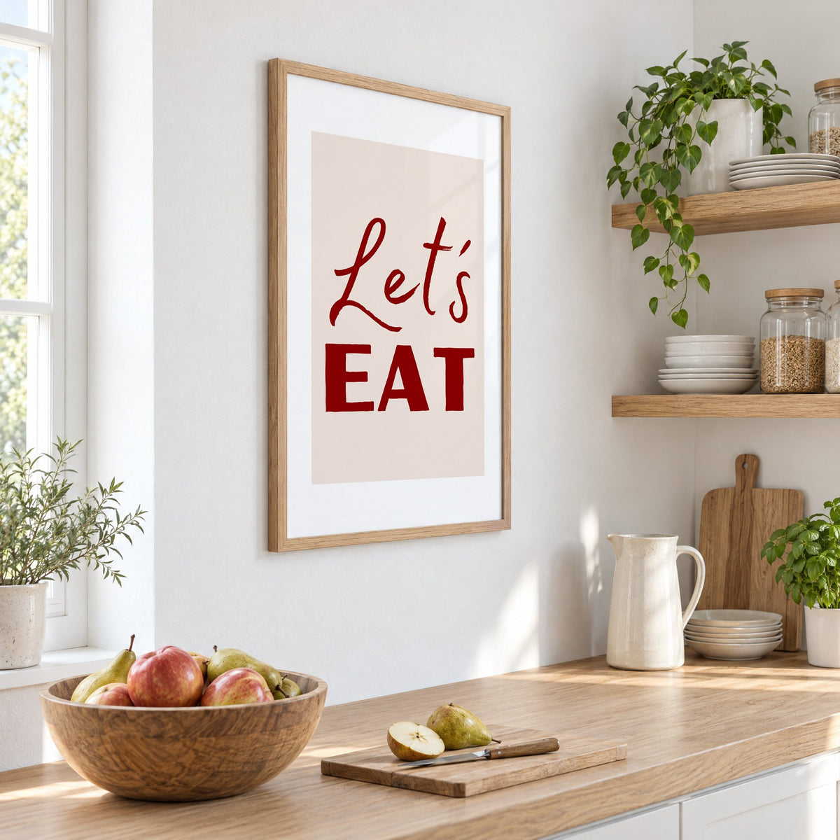 Let's EAT burgundy script print in an oak frame on a white kitchen wall above a counter with apples and pears, seen at an angle