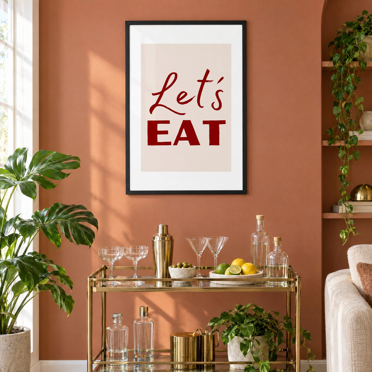 Let's EAT burgundy script print in a black frame on a terracotta wall above a brass bar cart with coupes and citrus