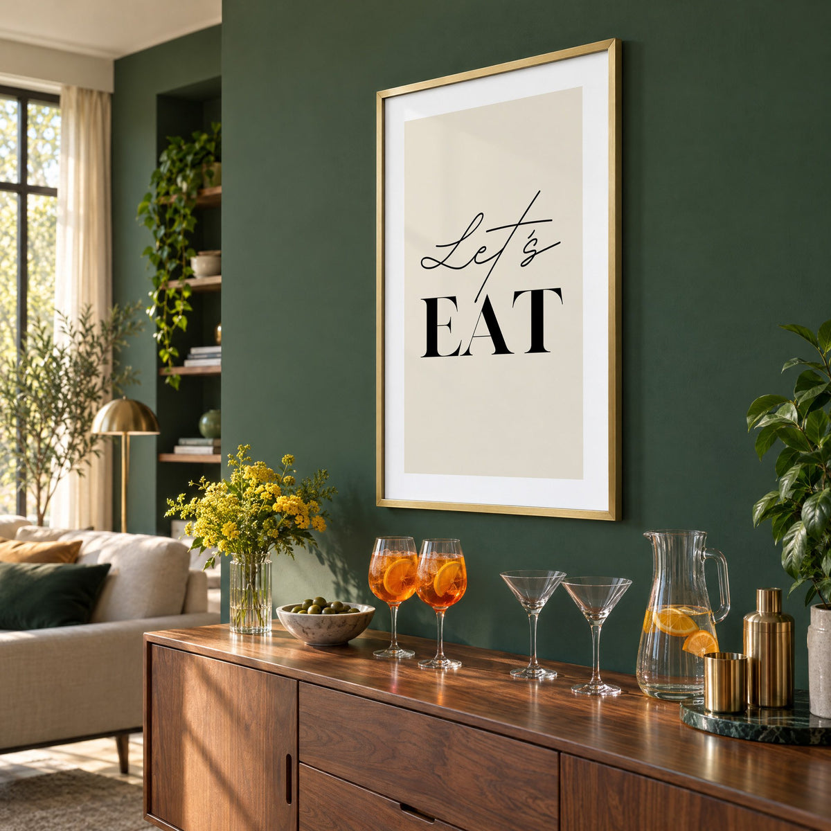 Let's EAT serif and script print in a brass frame on a bottle green wall above a sideboard with spritz and martini glasses, seen at an angle