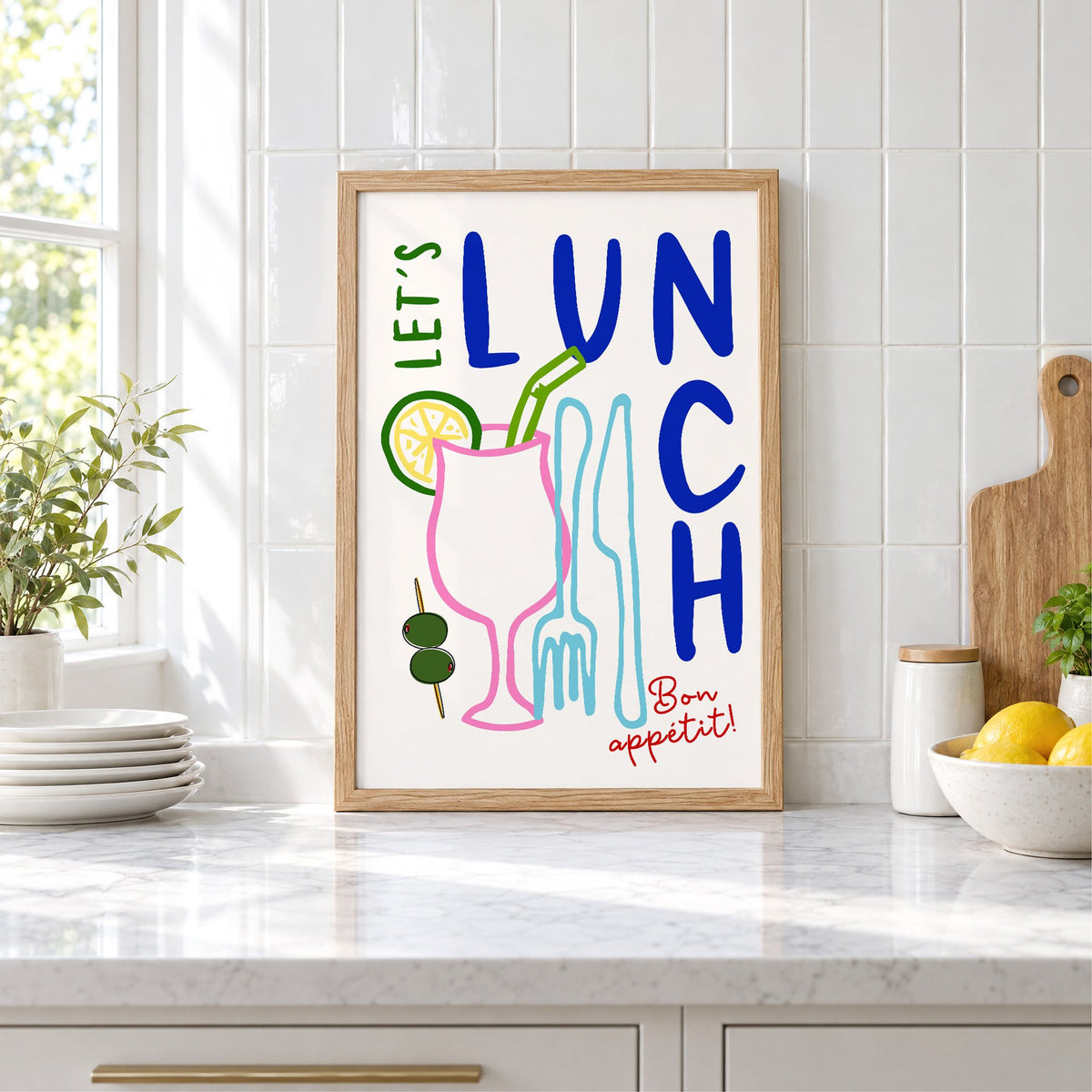 Let's Lunch cocktail and cutlery print in an oak frame leaning on a marble counter by white tiles and lemons