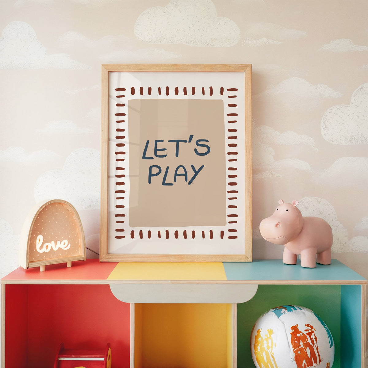 Lets play typography nursery print in an oak frame on a rainbow toy shelf against cloud wallpaper