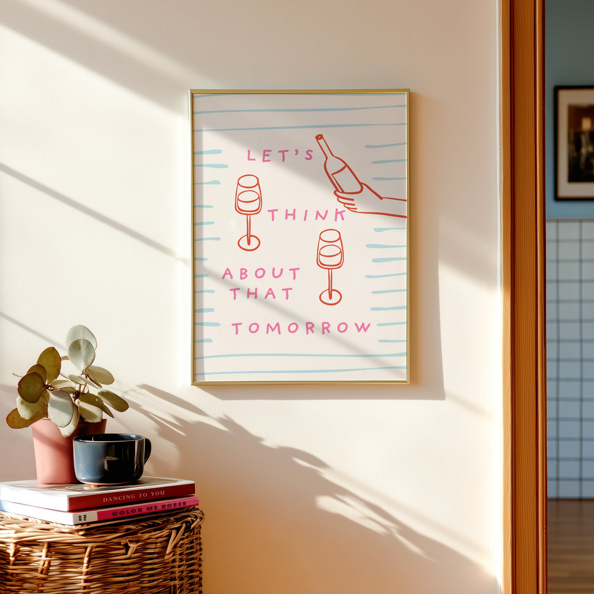 Wine pour line print shown in a gold frame above a basket of books and a pilea plant