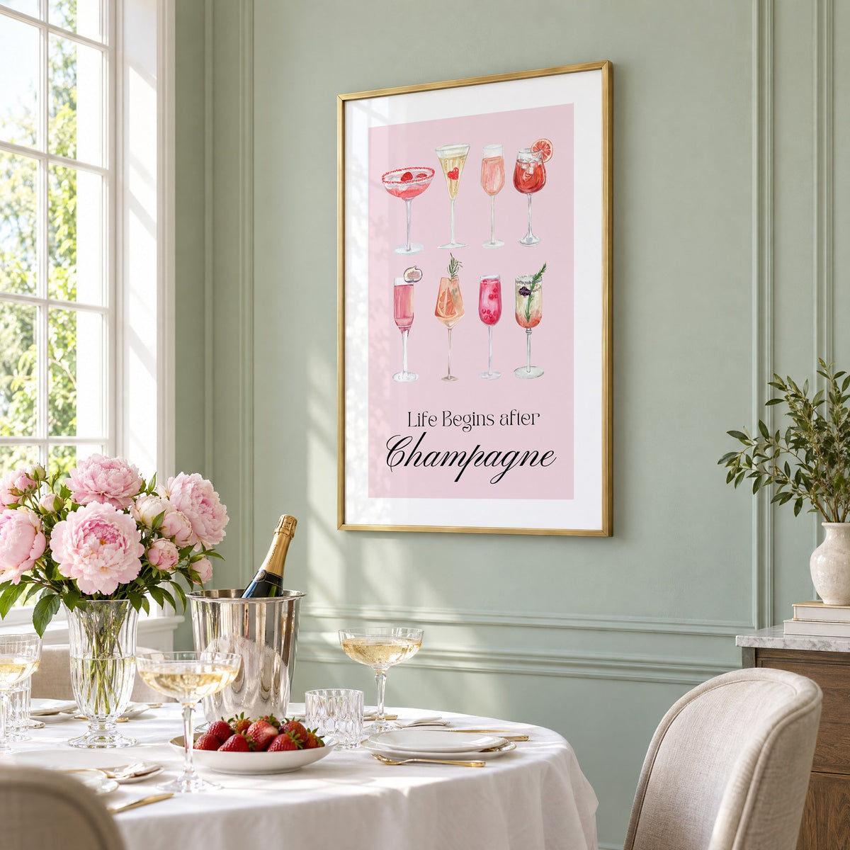 Life Begins after Champagne watercolor glasses print in a brass frame on a sage wall above a brunch table with champagne and peonies, seen at an angle