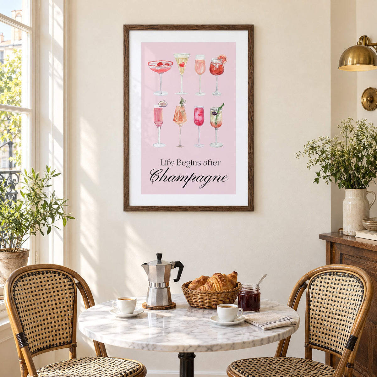 Life Begins after Champagne watercolor glasses print in a walnut frame above a marble bistro table with croissants and rattan chairs