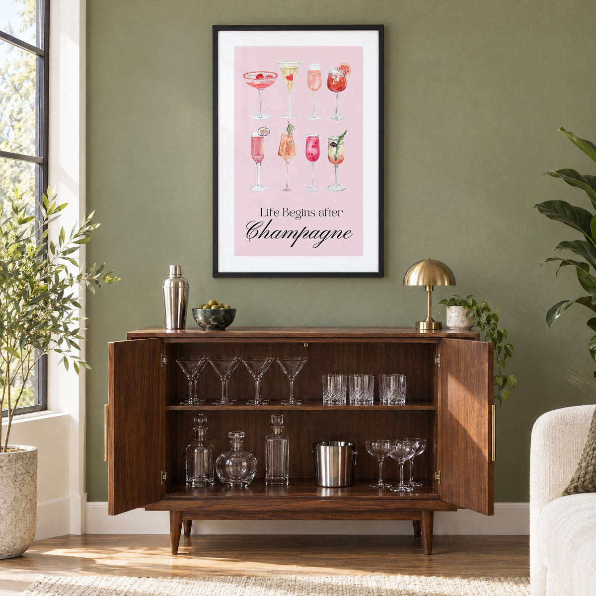 Life Begins after Champagne watercolor glasses print in a black frame on an olive green wall above a walnut bar cabinet with glassware