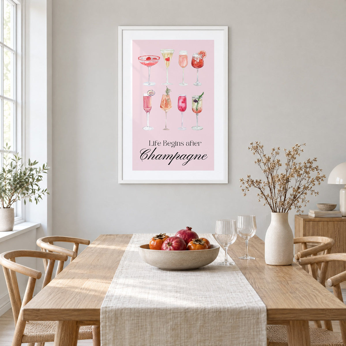 Life Begins after Champagne watercolor glasses print in a white frame above a light oak dining table with a linen runner and fruit bowl