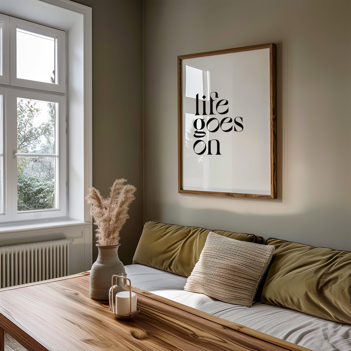 Life goes on serif quote print in a walnut frame above an olive daybed by a window