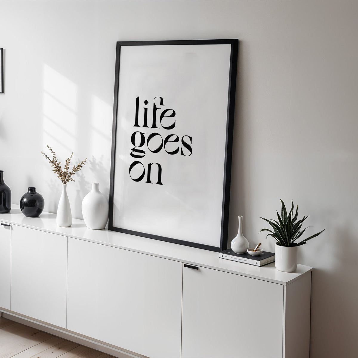 Life goes on serif quote print in a black frame on a white sideboard with black and white vases