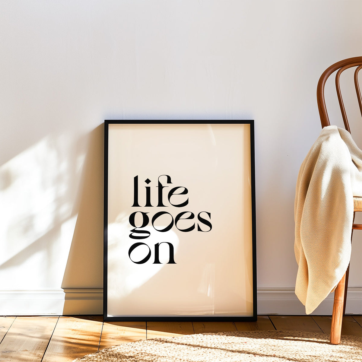 Life goes on serif quote print in a black frame leaning on a wood floor beside a bentwood chair
