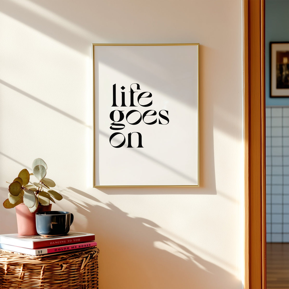 Life goes on serif quote print shown in a gold frame above a wicker basket and stacked books