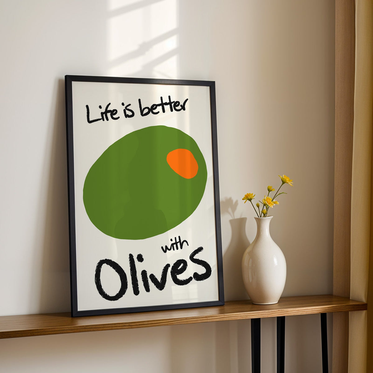 Life is better with olives print in a black frame on a console table with daisies