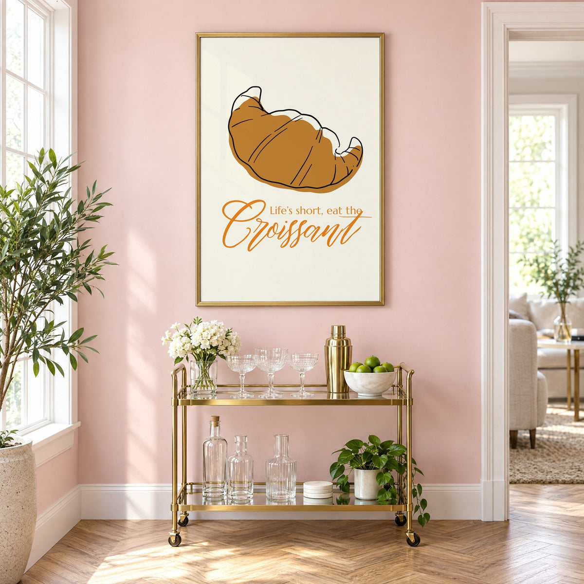 Life's short, eat the Croissant print in a brass frame on a blush pink wall above a gold bar cart with glassware