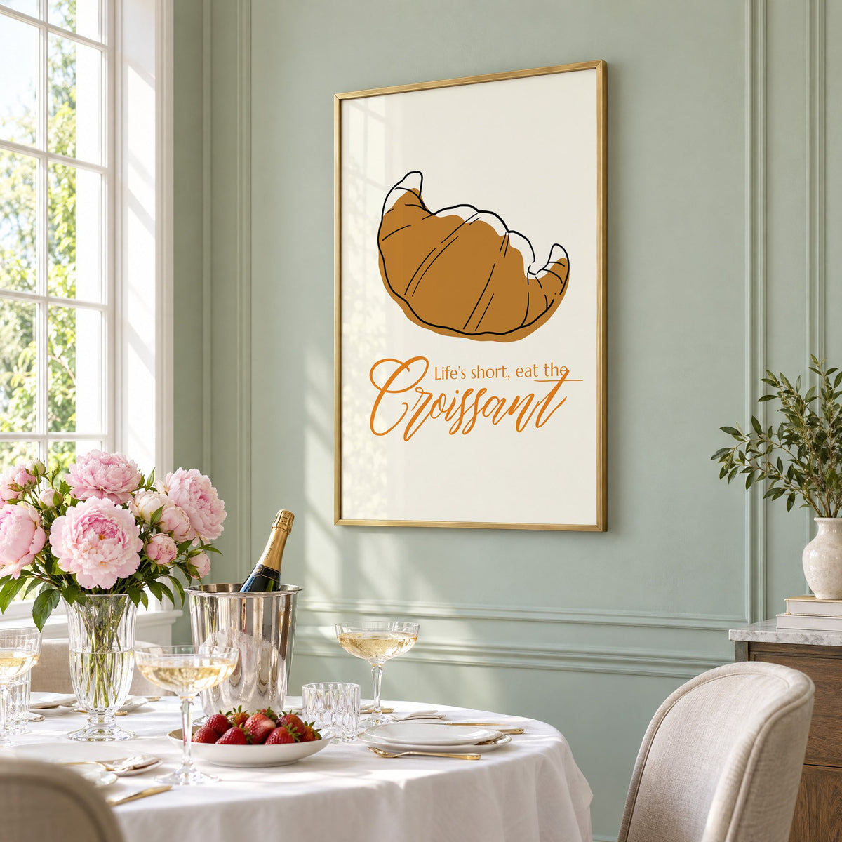 Life's short, eat the Croissant print in a brass frame on a sage wall above a brunch table with champagne and peonies, seen at an angle