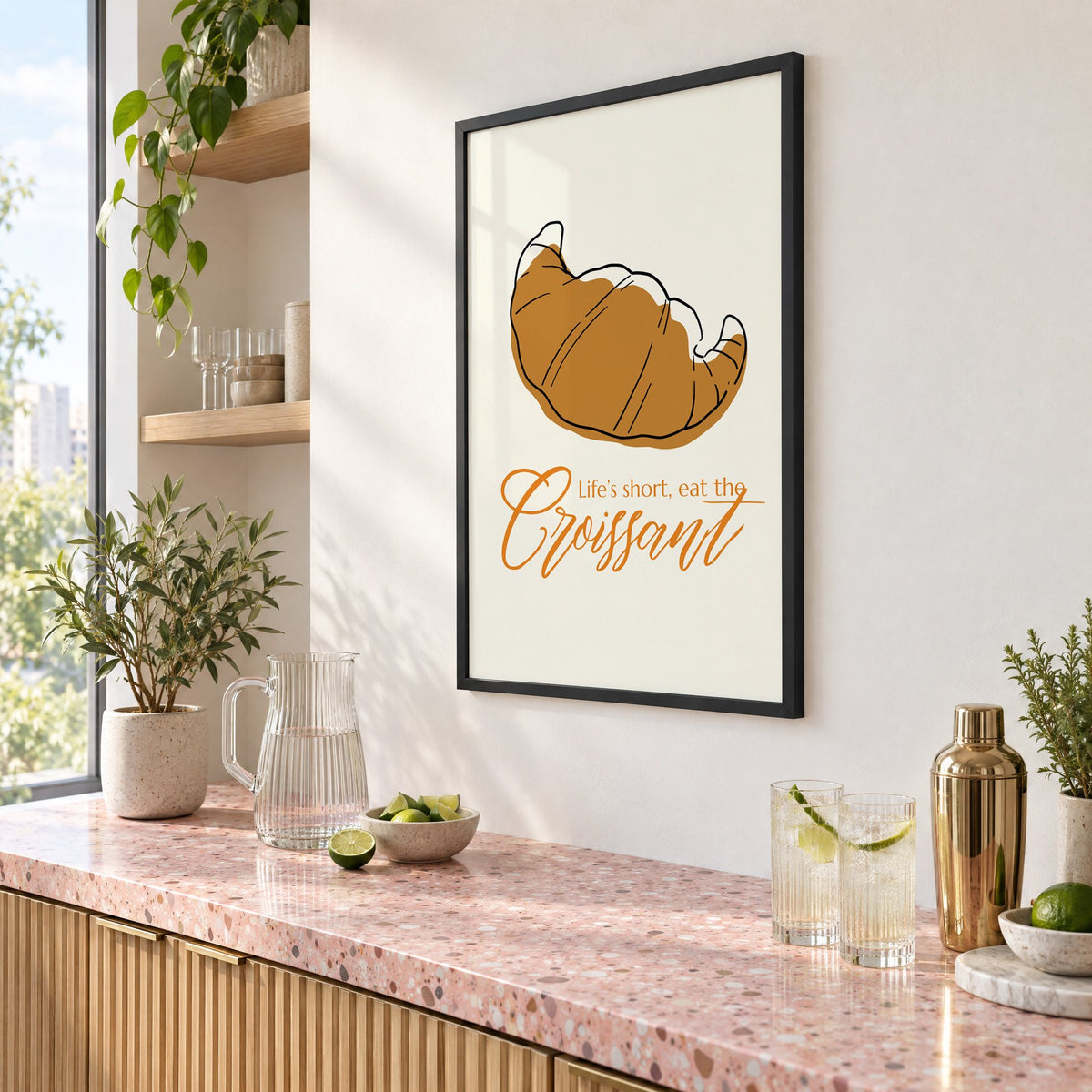 Life's short, eat the Croissant print in a black frame above a pink terrazzo cocktail counter with limes, seen at an angle